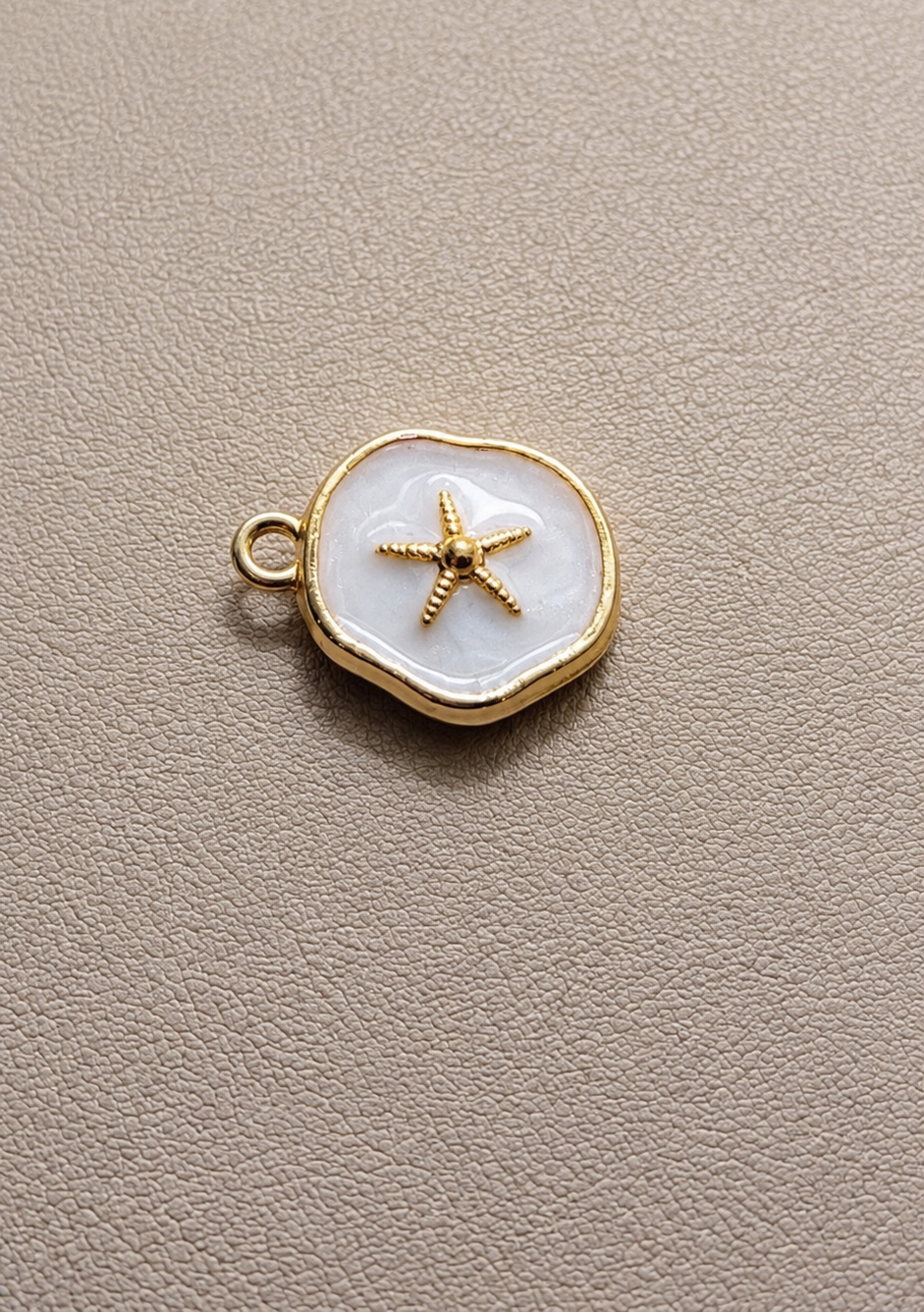 A small round white enamel charm with a gold border and a gold starfish in the center.